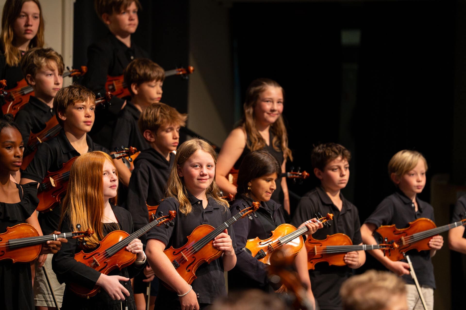 A group of children dressed in black shirts stand on stage holding violins, smiling and looking toward the audience, appearing to be at the end of a school musical performance.