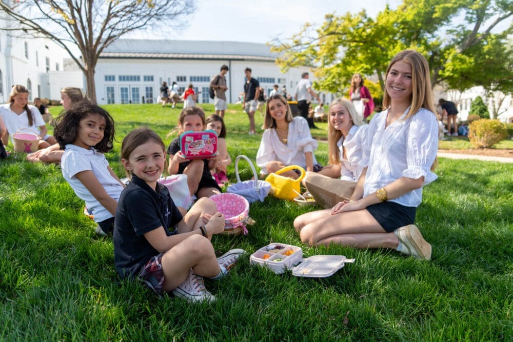 A group of smiling children and young adults sit on green grass having a picnic with lunch boxes and baskets, enjoying a sunny day outside a white school building.