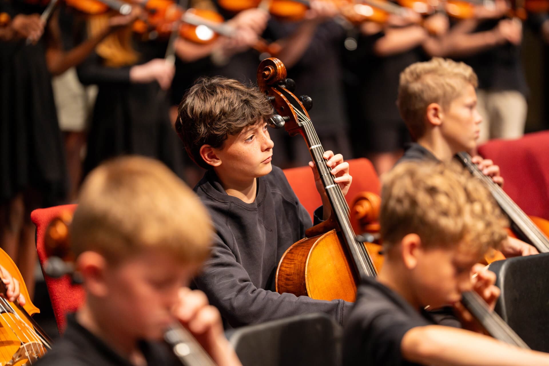 A group of young school musicians plays string instruments. In the foreground, a boy focuses on playing the cello, surrounded by other children and musicians holding violins, all wearing dark clothing.