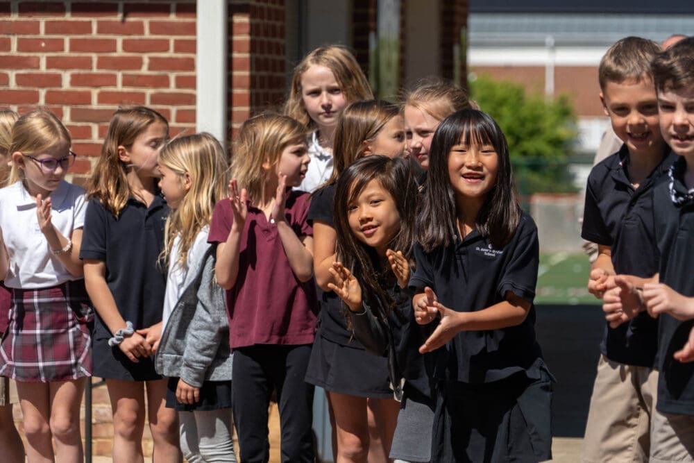 A group of school children in uniforms stand outside near a brick wall. Some are clapping and smiling, while others look on with curiosity. It is a sunny day.