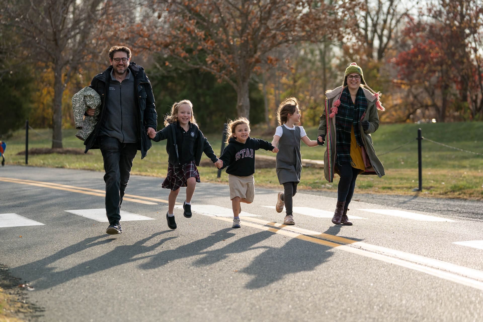 A family of five, two adults and three children, hold hands and joyfully walk or jump across a sunny road in a park, surrounded by autumn trees and fallen leaves.