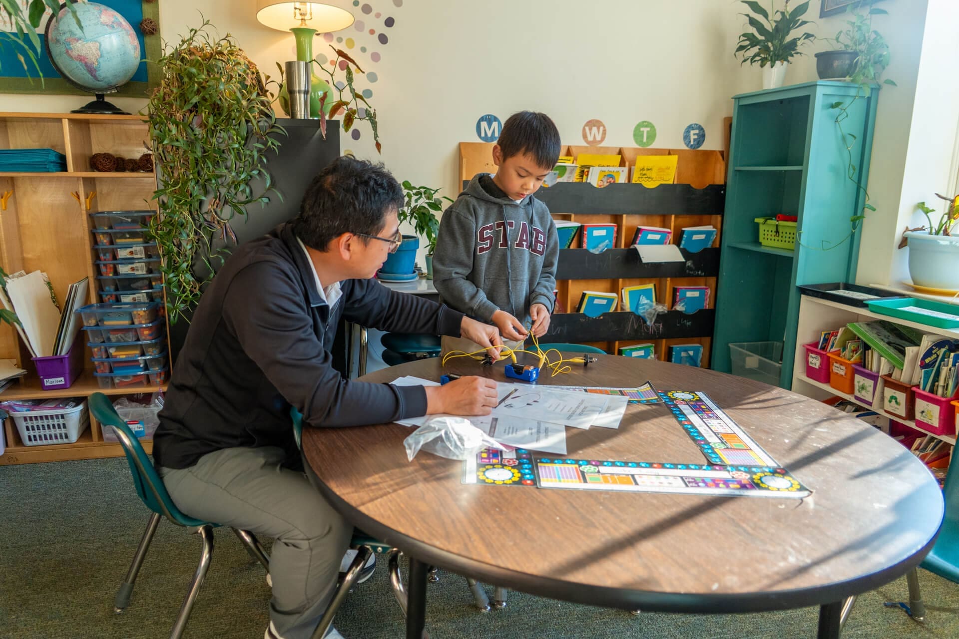A teacher sits at a round table helping a young student with a hands-on science project using wires. They are in a classroom with shelves, colorful bins, plants, and educational materials visible in the background.