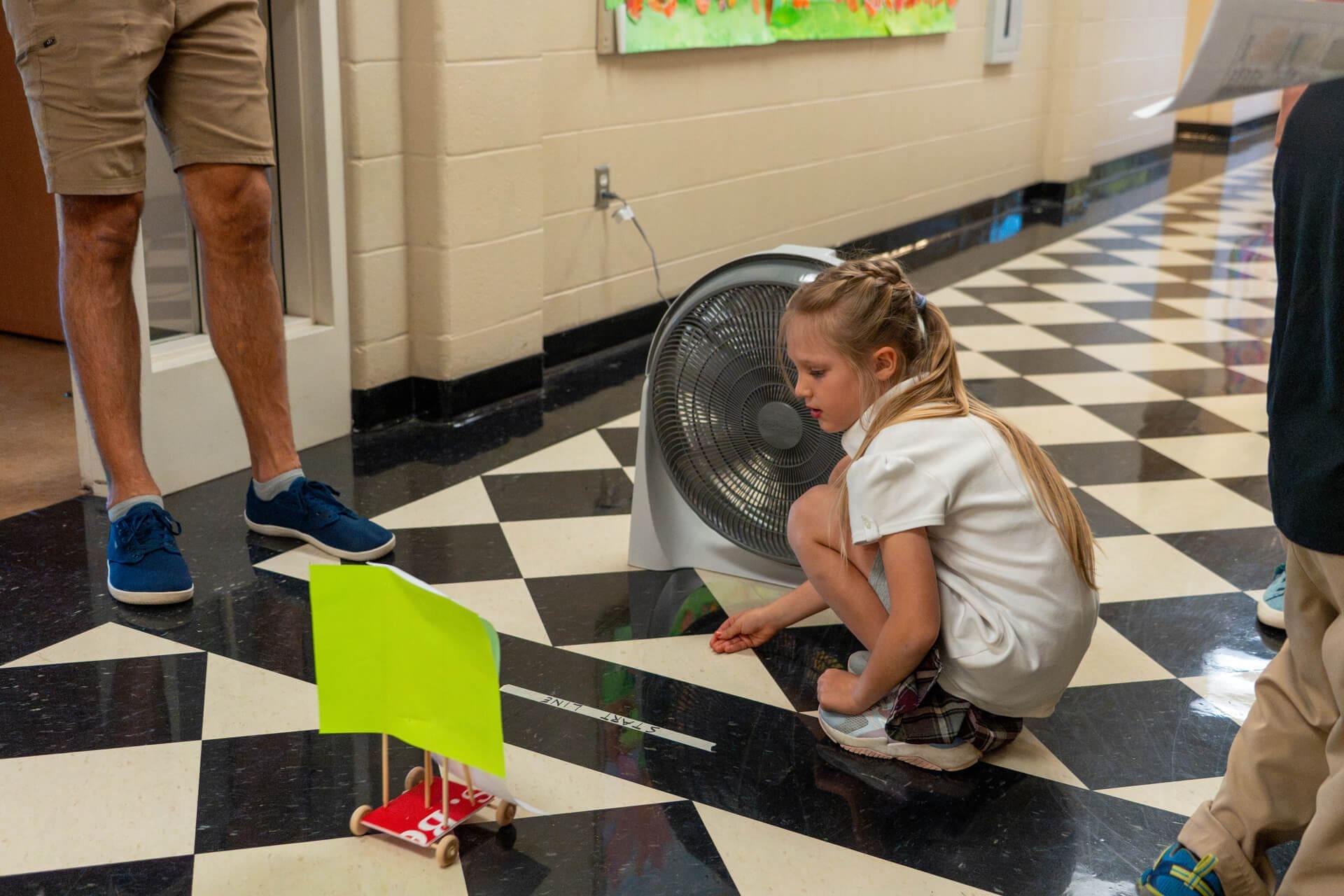 A young girl crouches on a checkered hallway floor, observing a small model car with a green sail positioned in front of a blowing fan while an adult stands nearby.