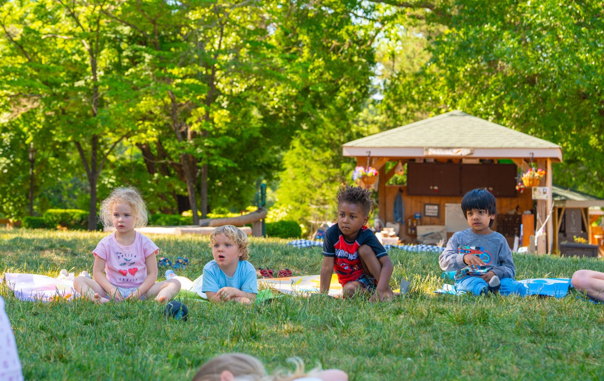 Four young children sit on grass outdoors on a sunny day, spaced out on blankets. Behind them is a small pavilion or gazebo with trees and greenery in the background.