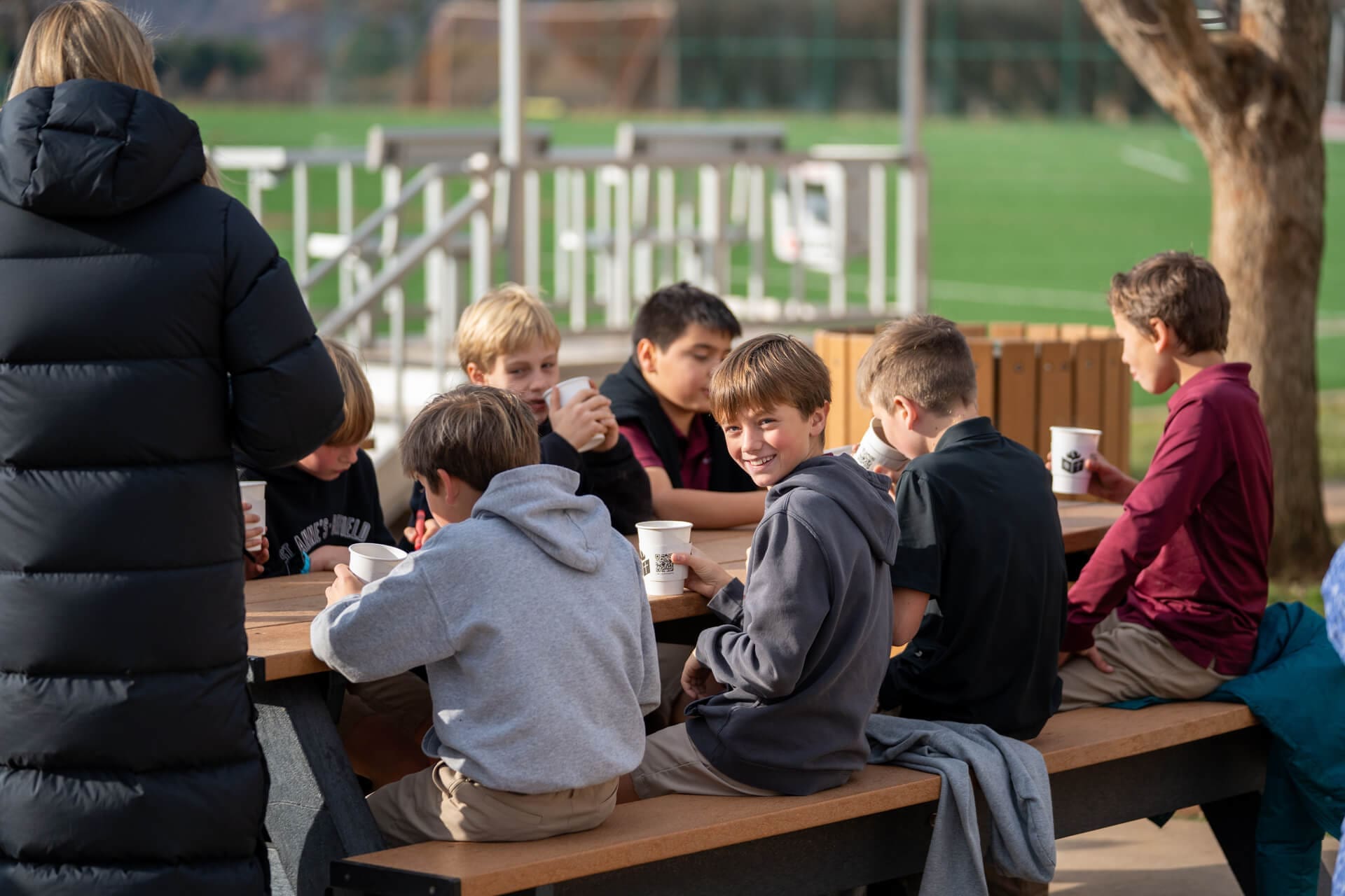 A group of boys sit at a picnic table outdoors, drinking from cups and talking, likely during a school outing. One boy in the center is smiling at the camera. A person in a black coat stands nearby, with a field and playground in the background.