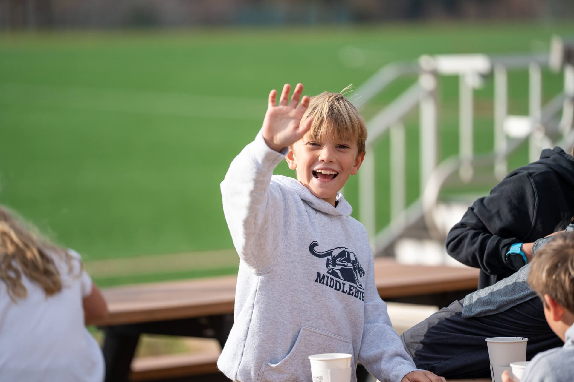 Smiling boy in a gray Middlebury hoodie waves at the camera while sitting at an outdoor table with other kids, with a green sports field in the background.