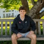 A young man with short brown hair, wearing a black Nike jacket and gray shorts, sits smiling on a wooden bench in a sunny park with green grass and a large tree behind him.
