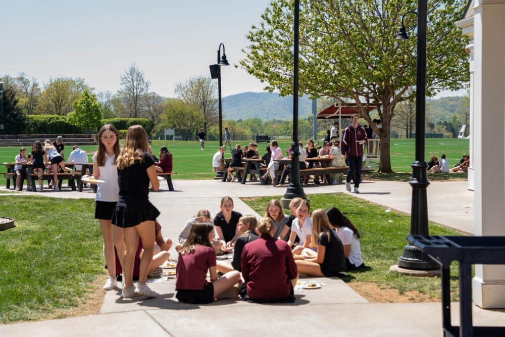 Students in uniforms sit and stand in groups eating lunch outside on a sunny day, near picnic tables and a grassy field, with trees and hills in the background.