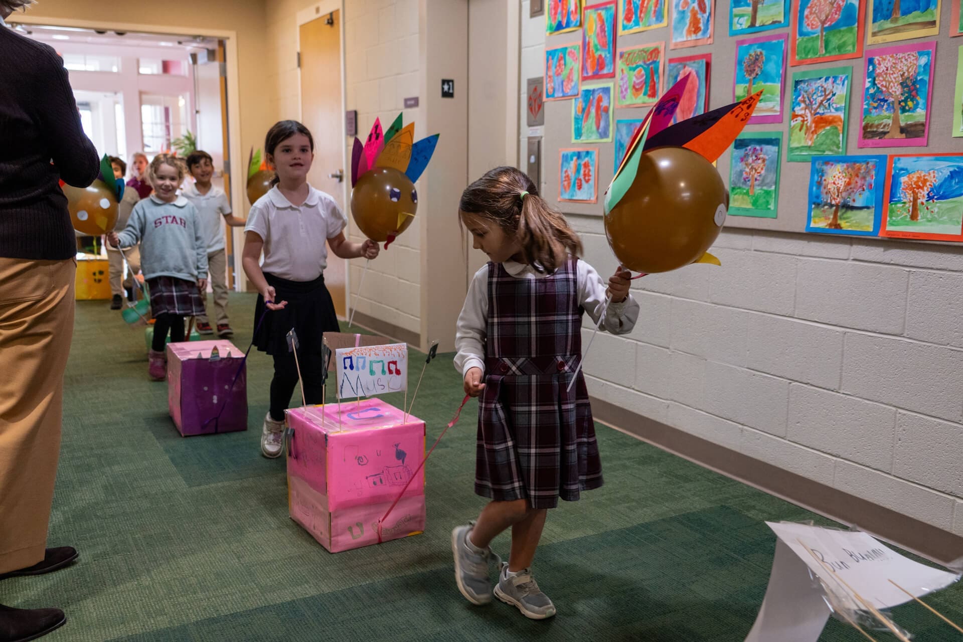 Young children walk in a school hallway pulling decorated boxes with turkey balloons, wearing uniforms. Colorful artwork is displayed on the wall, and a teacher is guiding them.