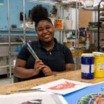 A smiling young woman sits at a table in an art studio, holding paintbrushes. Paint cans, colorful artwork, and art supplies are on the table and shelves are visible in the background.