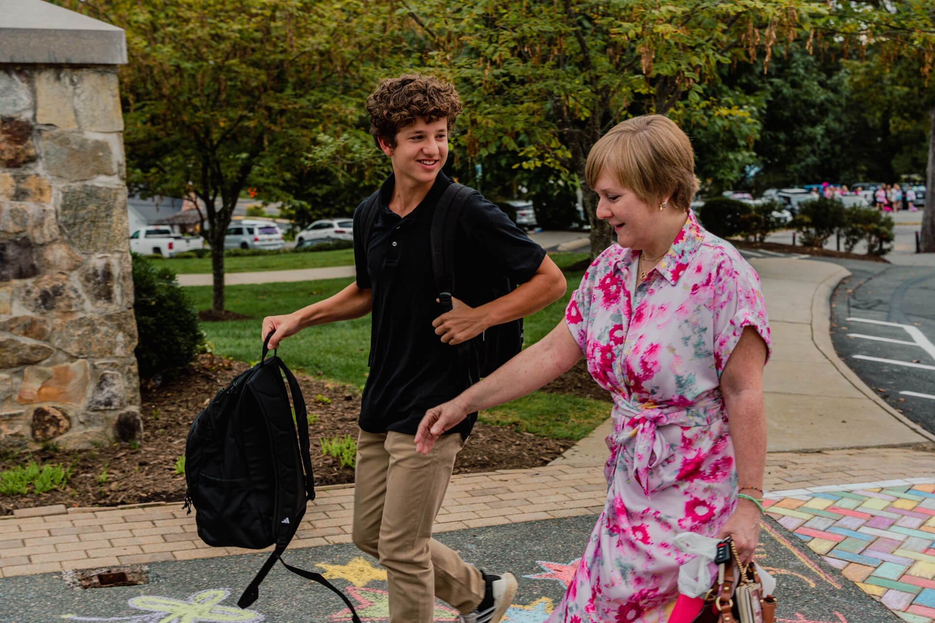 A teenage boy with a backpack and a woman in a pink floral dress walk together outside near a mural with colorful chalk art and a stone wall, smiling and talking. Trees and parked cars are in the background.