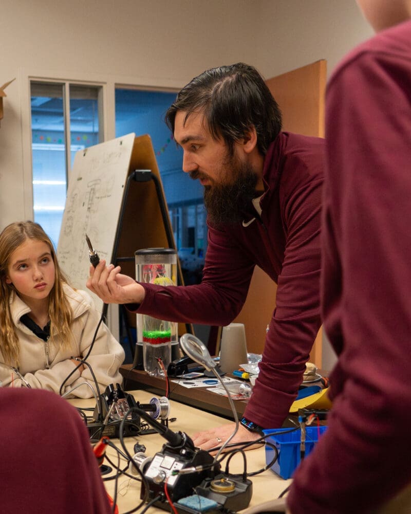 A bearded man leans over a table with electronics, holding a component and explaining to students; a girl listens attentively. A whiteboard with diagrams and various tools are visible in the background.