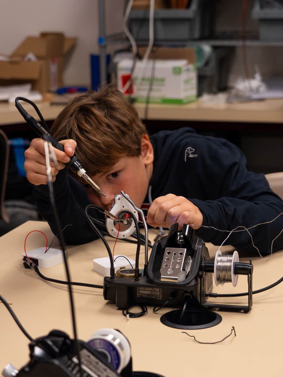 A boy carefully uses a soldering iron to work on an electronic project at a table, surrounded by wires, tools, and electronic components in a workshop setting.
