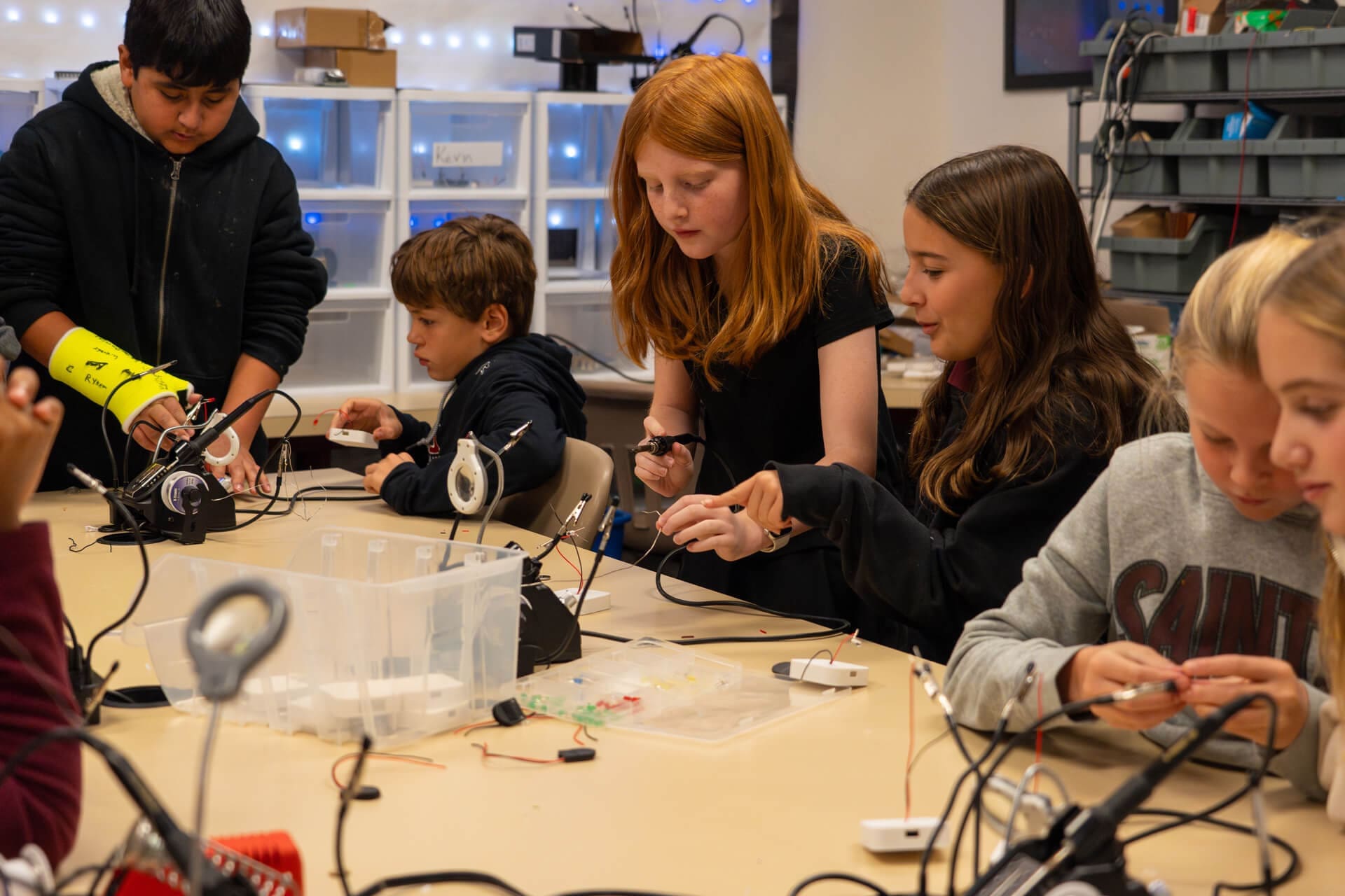 A group of students sits around a table working with wires and electronic components in a classroom, focused on a hands-on STEM project. Storage bins and tools are visible in the background.