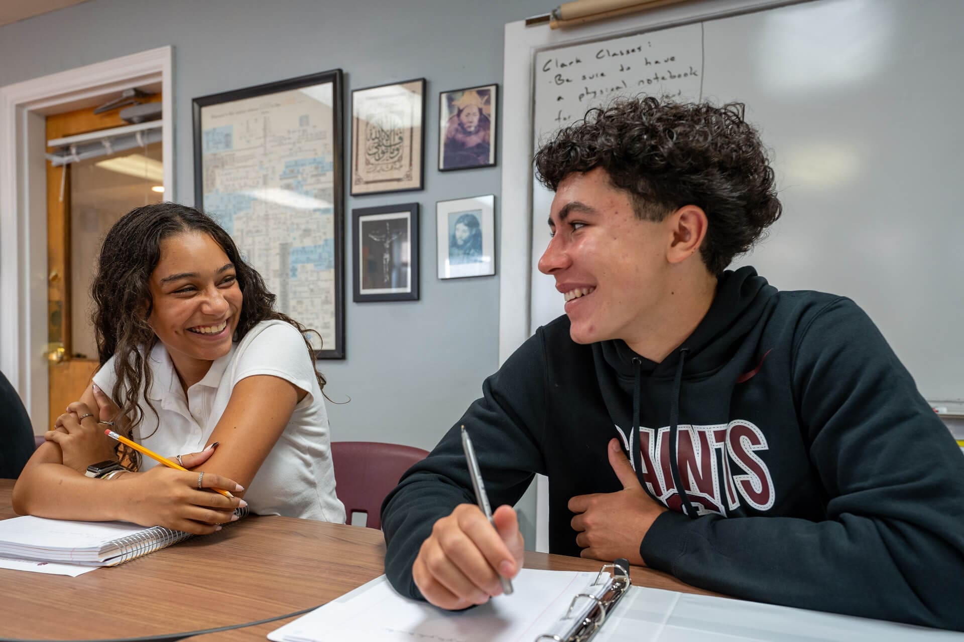 Two students sit at a classroom table, smiling and talking to each other while holding pencils and notebooks. Behind them, a whiteboard, framed photos, and a map hang on the wall.