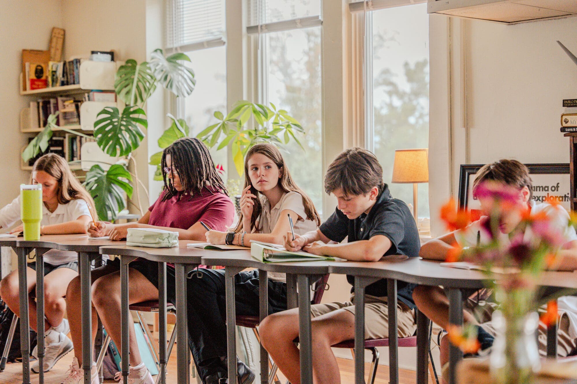 A group of students sit at desks arranged in a semi-circle in a bright classroom, writing in notebooks. There are plants and books on shelves in the background and a vase of flowers in the foreground.