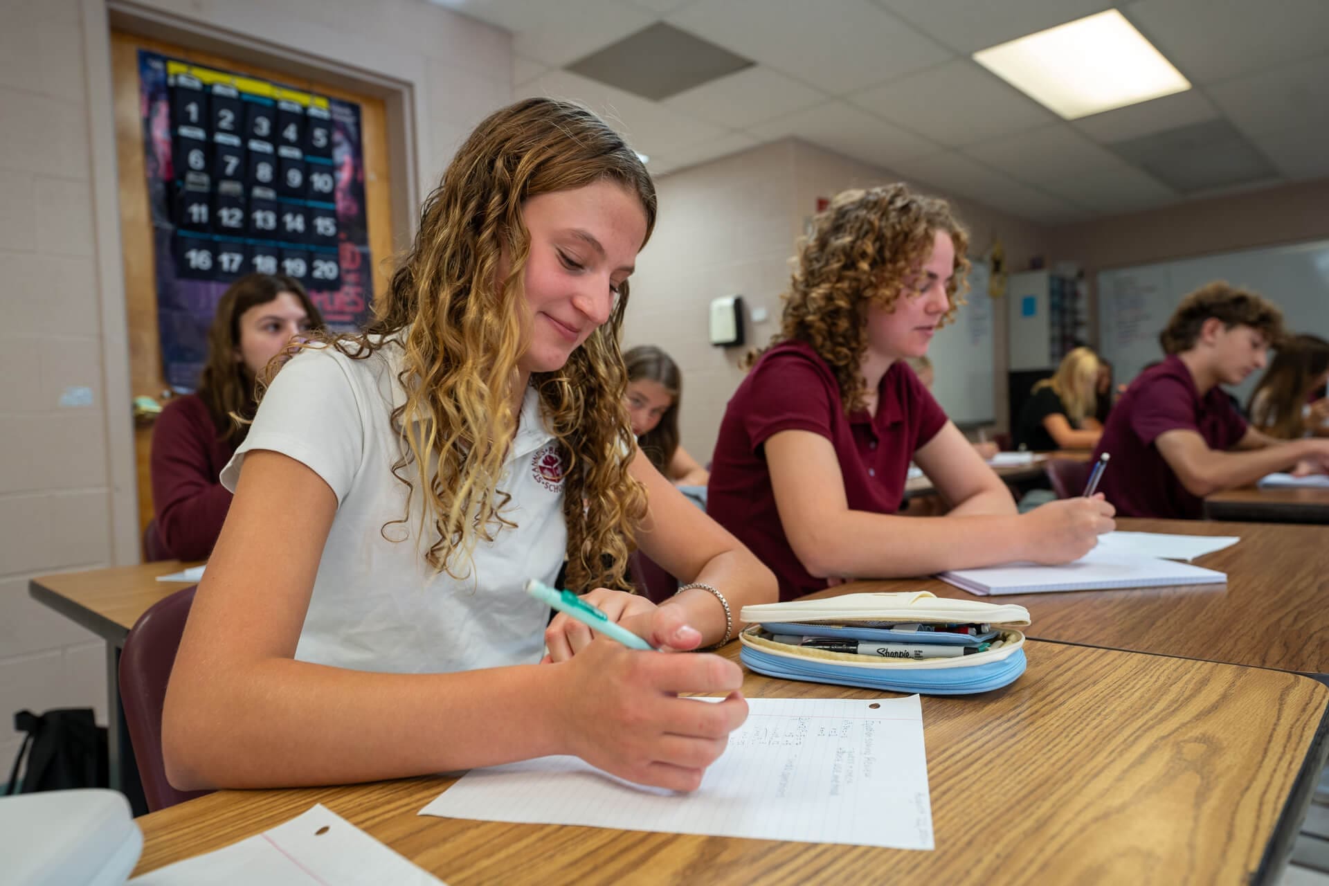 Several high school students sit at desks in a classroom, writing in notebooks. A girl in the foreground with curly hair and a white shirt smiles as she writes. Other students are focused on their work.