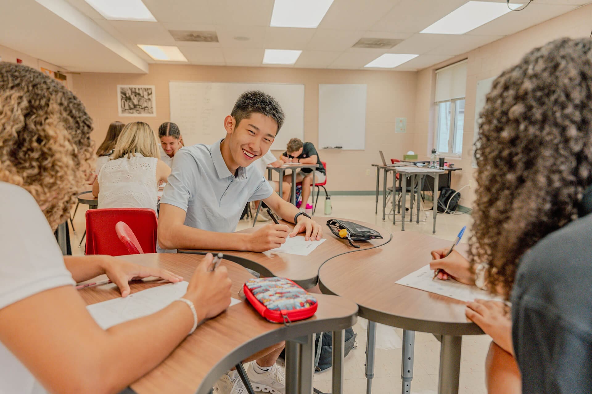 A group of high school students sit around a table in a bright classroom, writing on papers and smiling as they work together. Other students are seen in the background at separate desks.