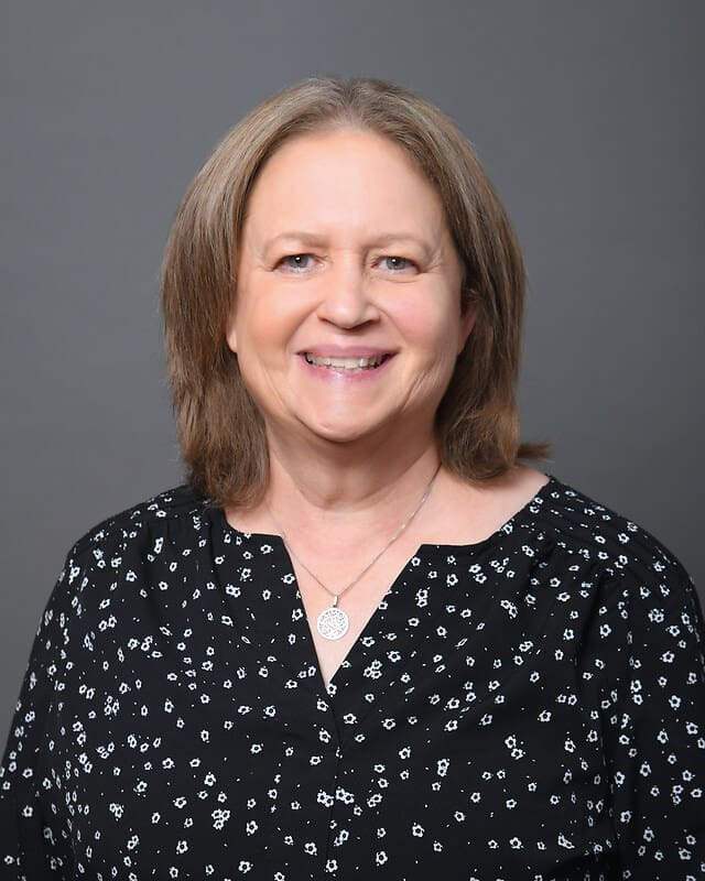 A woman with shoulder-length light brown hair, wearing a black blouse with white floral patterns and a circular pendant necklace, smiles in front of a plain gray background.