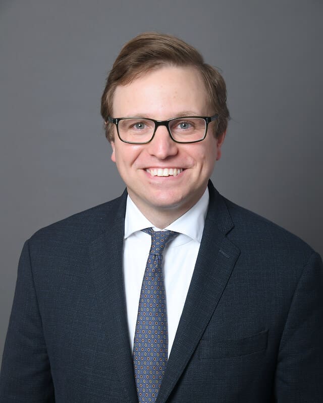 A man with light brown hair and glasses, wearing a dark suit, white shirt, and patterned tie, smiles at the camera against a plain gray background.