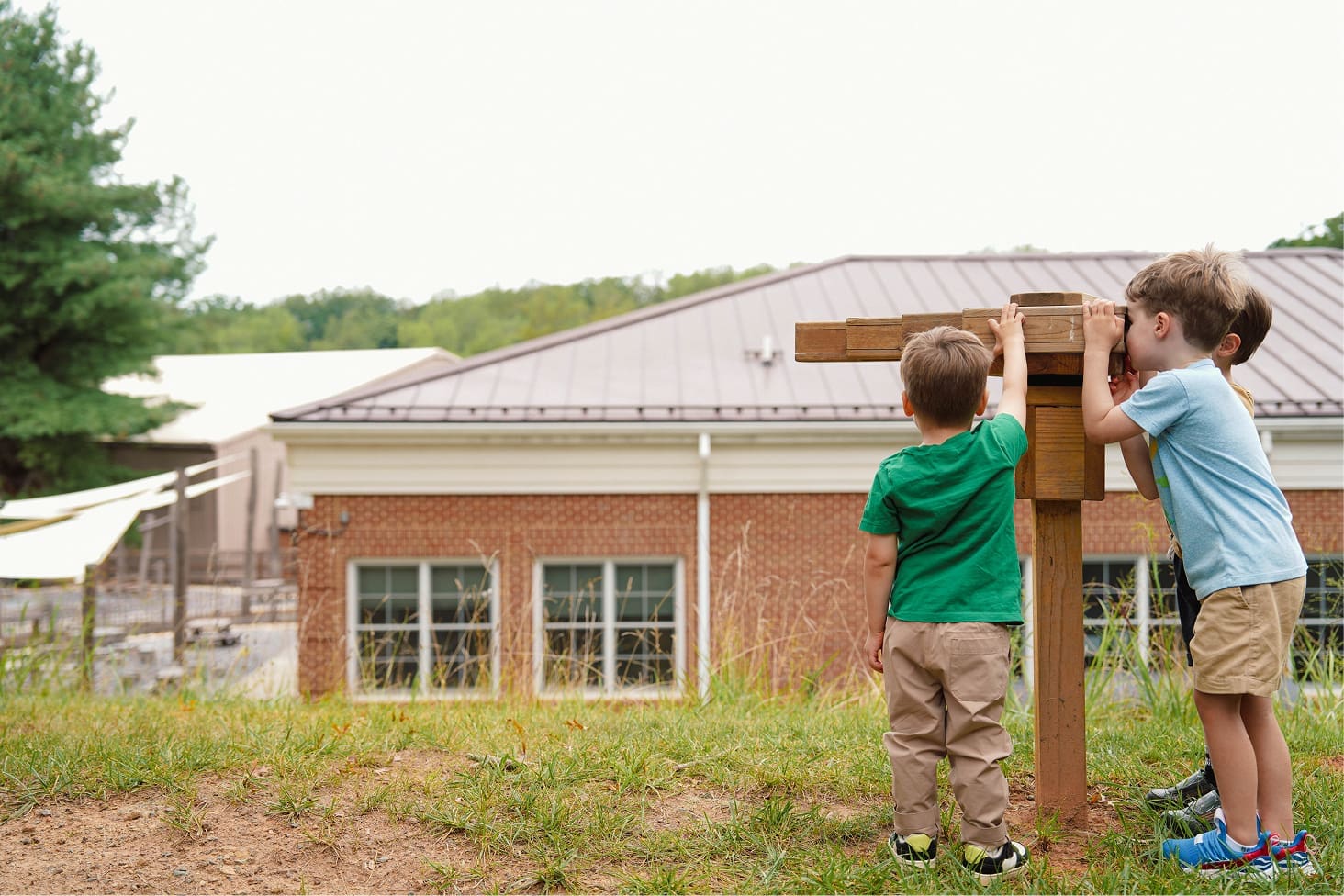 Three young boys stand on grass, with two looking through a wooden structure resembling binoculars, facing a brick building with large windows in the background. Trees and greenery are visible around the building.