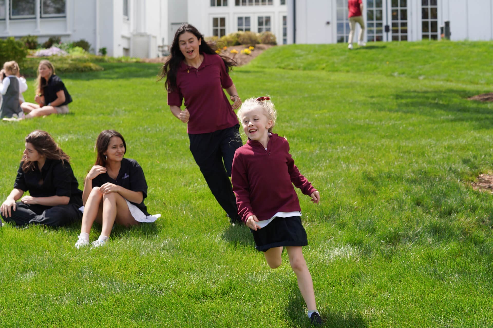 A young girl runs across a green lawn, smiling, while an adult follows behind her. Two other people sit on the grass nearby, and a white building is in the background.