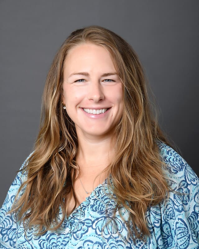 A woman with long, wavy light brown hair smiles at the camera. She is wearing a blue patterned blouse and standing in front of a plain gray background.