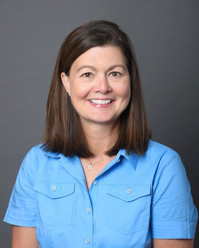 A woman with straight brown hair wearing a light blue short-sleeve button-up shirt smiles at the camera against a plain gray background.