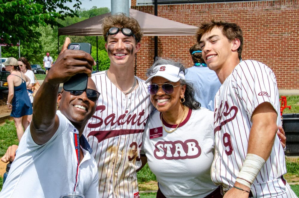 Four people, including two baseball players in striped uniforms reading Saints, pose for a smiling selfie outdoors. A woman in a STAB shirt and hat stands in the middle. Others and trees are visible in the background.