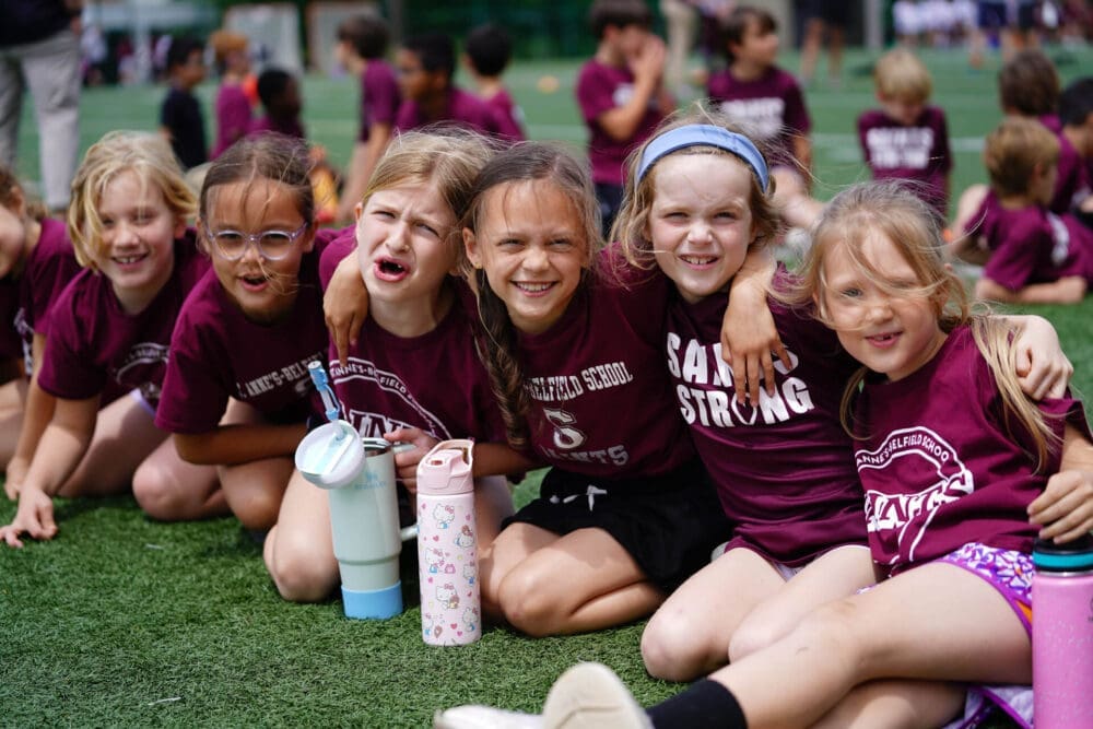 A group of young children in matching maroon shirts sit and smile together on a grassy field, arms around each other, with water bottles in front of them and other kids in the background.