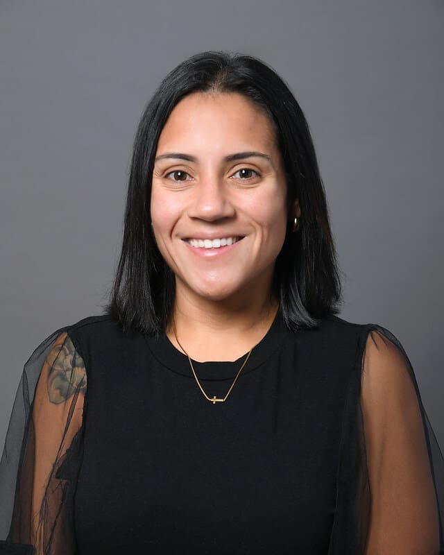 A woman with straight, shoulder-length black hair is smiling at the camera. She is wearing a black top with sheer sleeves, a gold necklace, and has a tattoo on her upper left arm. The background is plain gray.