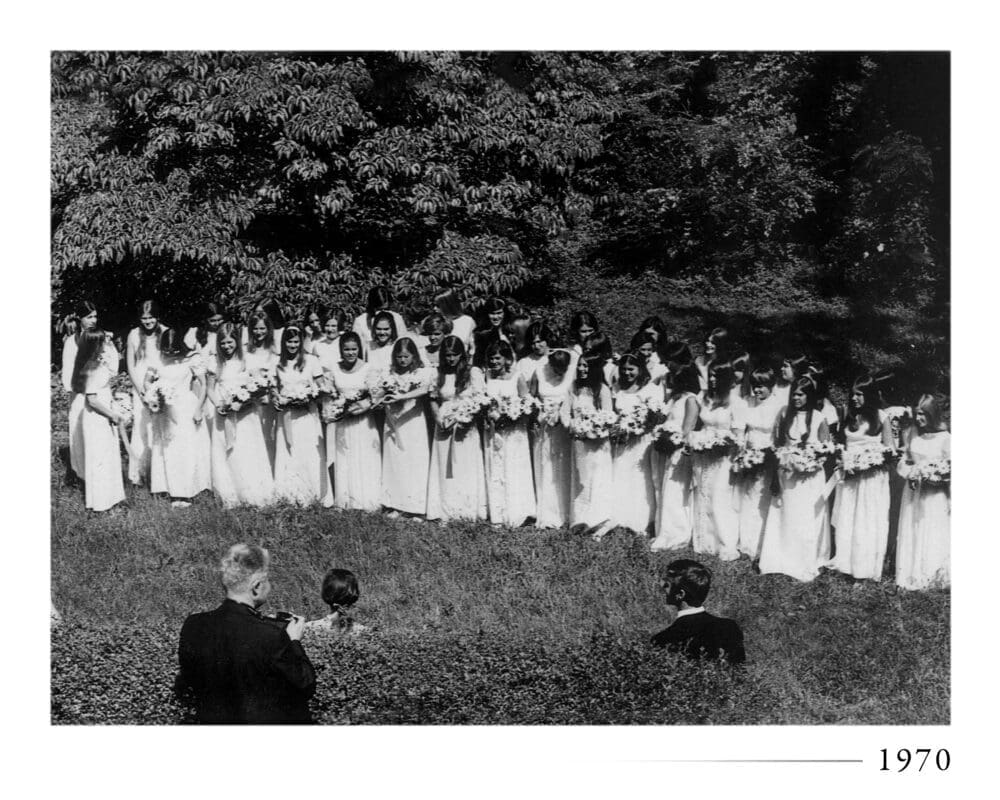 A group of women in white dresses holding bouquets stand in a grassy outdoor area while a few people observe and take photos. Trees fill the background. The photo is black and white and labeled 1970 in the corner.