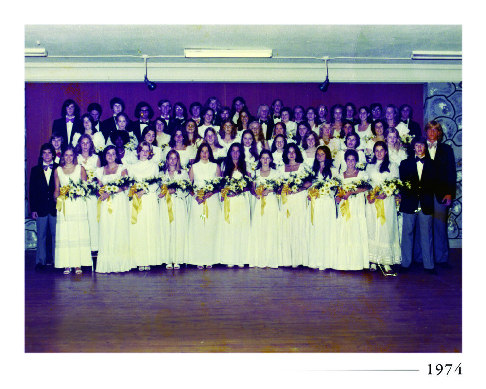 A large group of young men in tuxedos and young women in white dresses holding bouquets pose together on a stage for a formal group photo. The year 1974 is printed in the bottom right corner.
