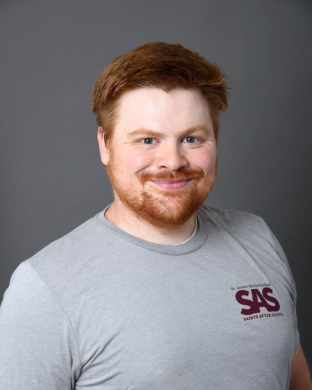A smiling man with short red hair and a beard, wearing a light gray t-shirt with a maroon SAS logo and text on the chest, stands against a plain gray background.