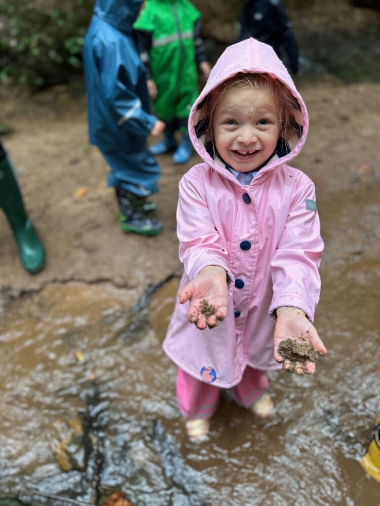 Sabrina in a pink raincoat smiles and holds out muddy sand in both hands while standing in shallow water. Other children in colorful raincoats are visible in the background.