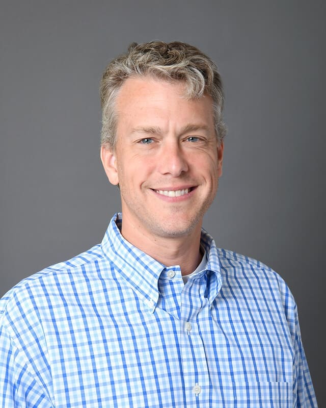 A man with short, curly gray hair and light skin smiles at the camera. He is wearing a blue and white plaid button-up shirt and stands in front of a plain gray background.