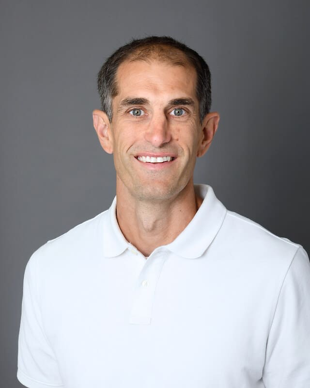 A man with short brown hair and light eyes is smiling at the camera. He is wearing a white polo shirt and standing in front of a plain gray background.