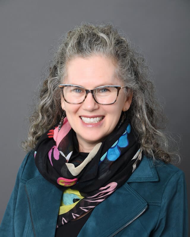 A woman with curly gray hair and glasses smiles at the camera. She is wearing a teal jacket and a black scarf with colorful patterns, set against a plain gray background.