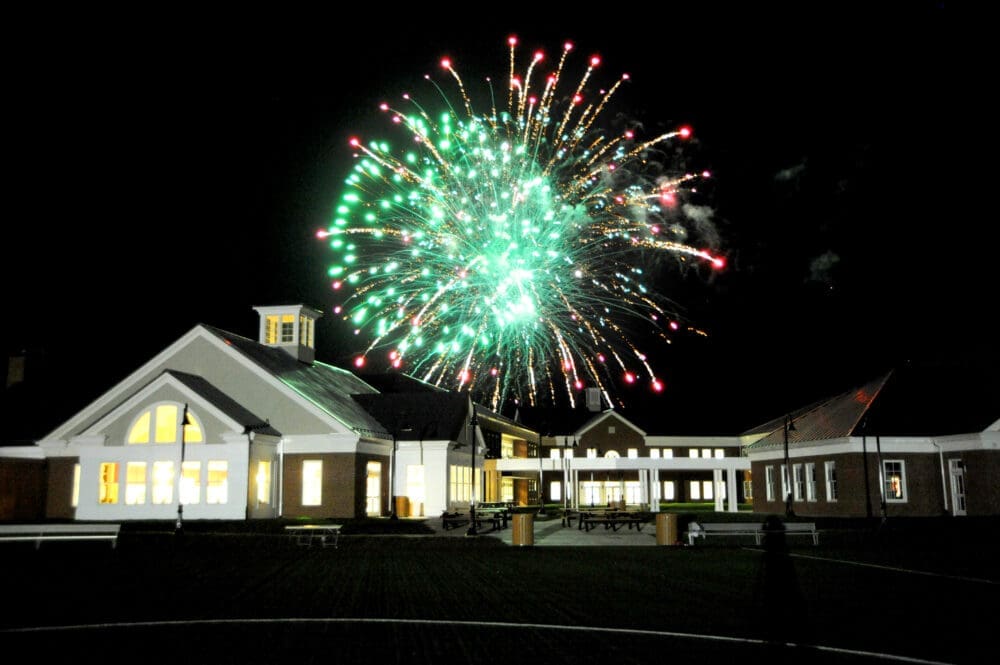 Bright green and red fireworks light up the night sky above a large, well-lit building with a pointed roof and cupola, surrounded by smaller connected structures and an open courtyard.