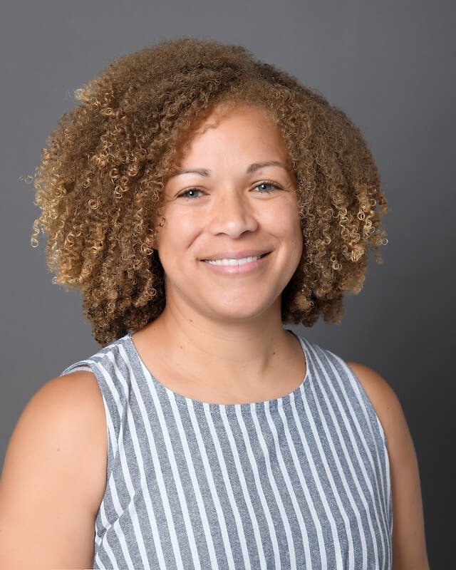 A woman with curly light brown hair smiles at the camera. She is wearing a sleeveless, gray and white striped top and stands in front of a plain gray background.