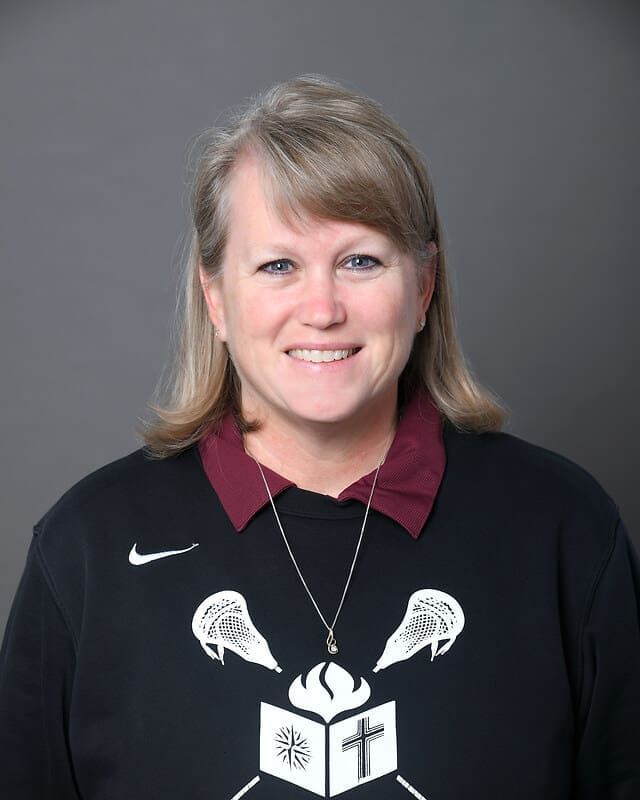 A woman with light brown hair and side-swept bangs smiles at the camera. She is wearing a black sweatshirt with a logo, a maroon collared shirt underneath, and a necklace. The background is plain gray.