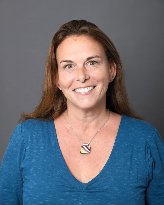 A woman with long brown hair wearing a blue V-neck top and a colorful square pendant necklace smiles in front of a plain gray background.