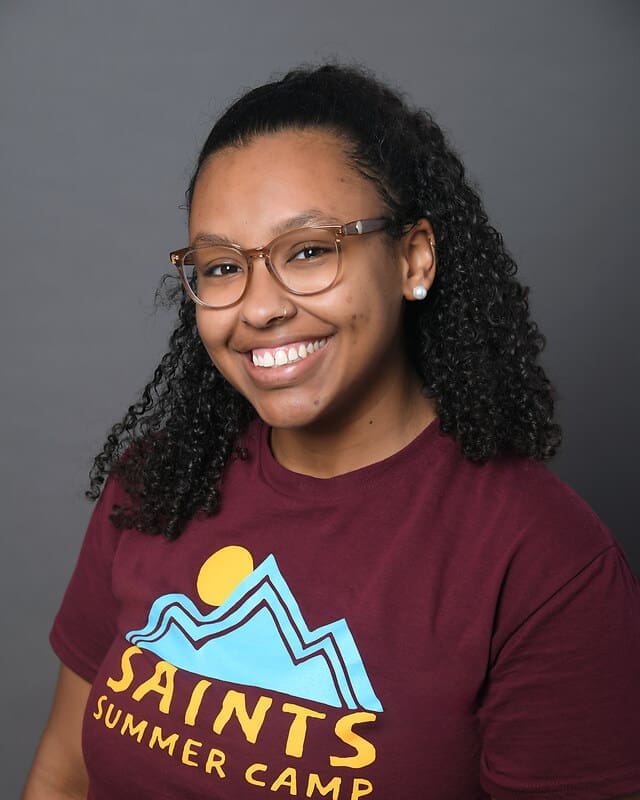 A smiling young woman with curly hair, wearing glasses and a maroon Saints Summer Camp T-shirt with a mountain and sun graphic, poses in front of a plain gray background.