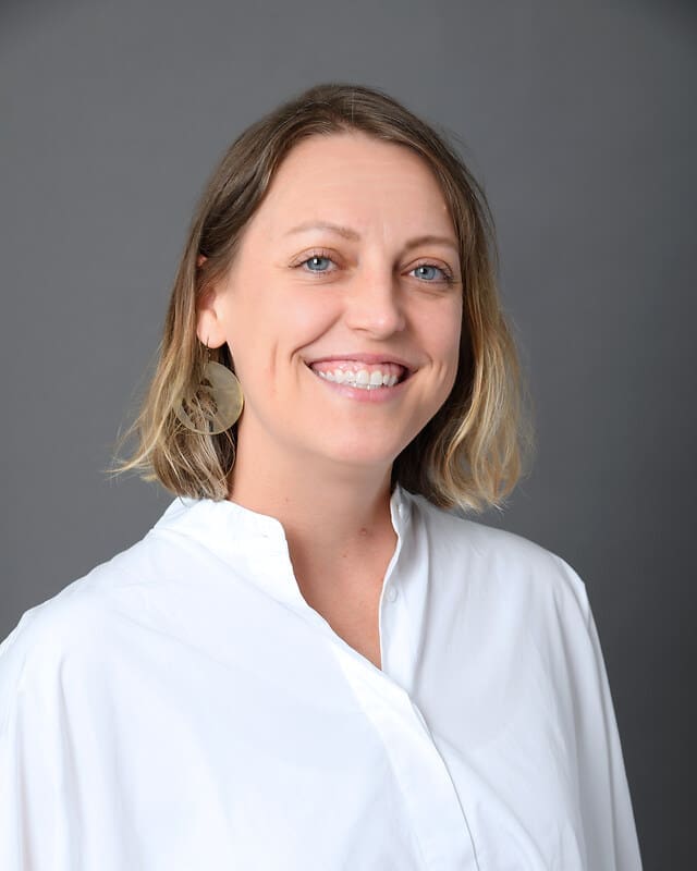 A smiling woman with shoulder-length blonde hair wears a white blouse and large round earrings, posing in front of a plain gray background.