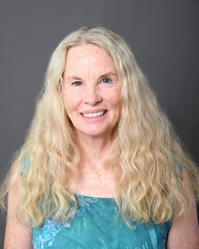 Smiling older woman with long, wavy blonde hair wearing a sleeveless blue patterned top, posing in front of a plain gray background.