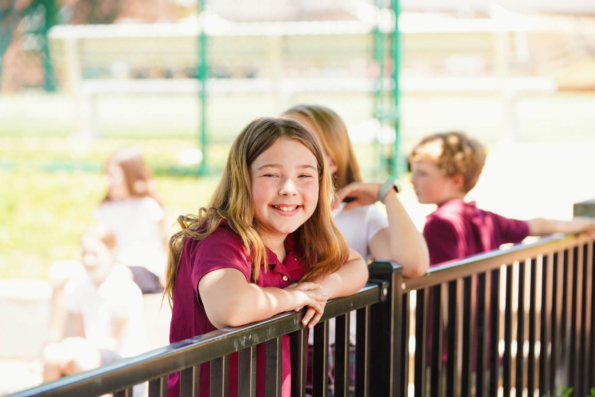 Smiling girl with long hair leans on a black metal fence, wearing a maroon shirt. Other children and playground equipment are blurred in the sunny background.