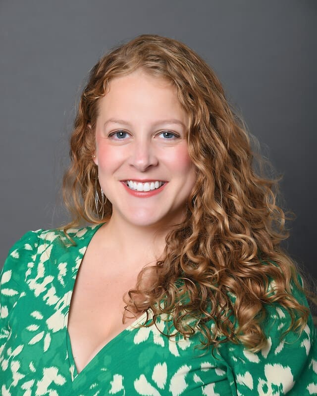 A woman with long, curly blonde hair smiles at the camera. She is wearing a green patterned dress with a V-neck and has hoop earrings. The background is plain gray.