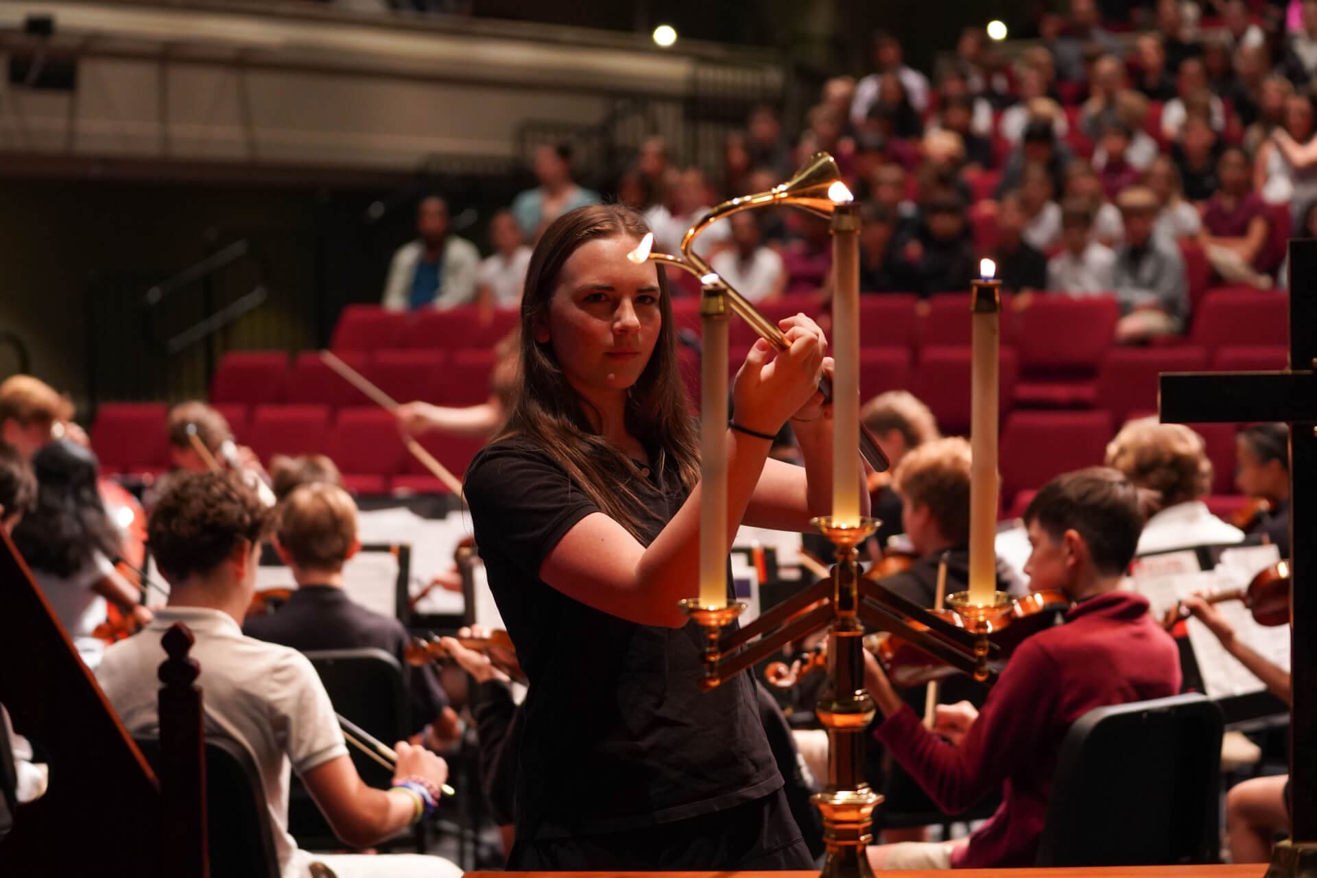 A person lights a large candelabrum on stage while an orchestra performs and an audience watches from red theater seats in the background.