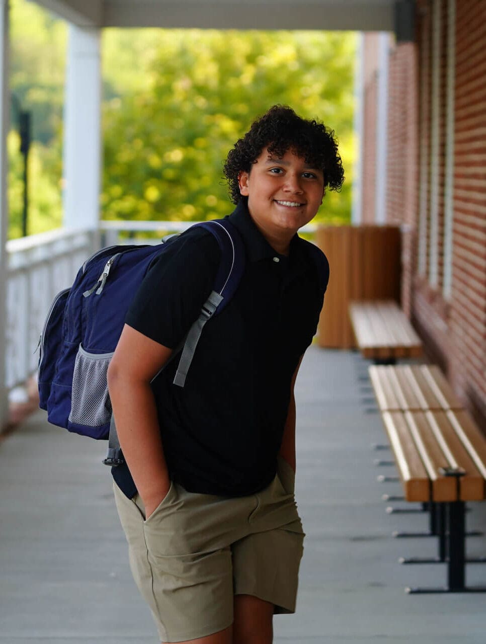 A smiling teen with curly hair, wearing a black polo shirt and khaki shorts, stands outdoors with a blue backpack on, next to benches and a brick building. Green trees are visible in the background.