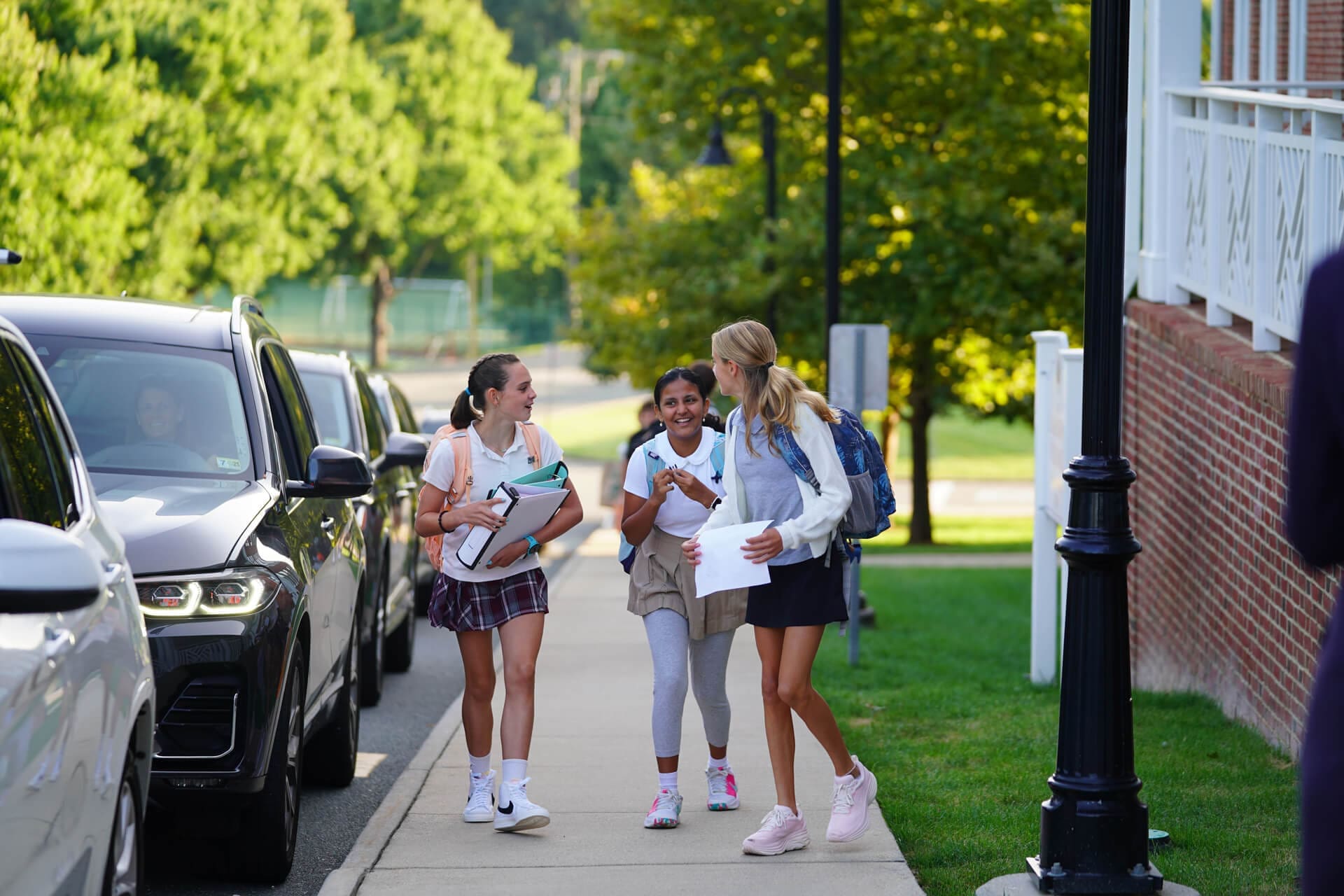 Three schoolgirls walk and chat happily on a sidewalk near parked cars and a brick building, carrying backpacks and folders, with green trees in the background.
